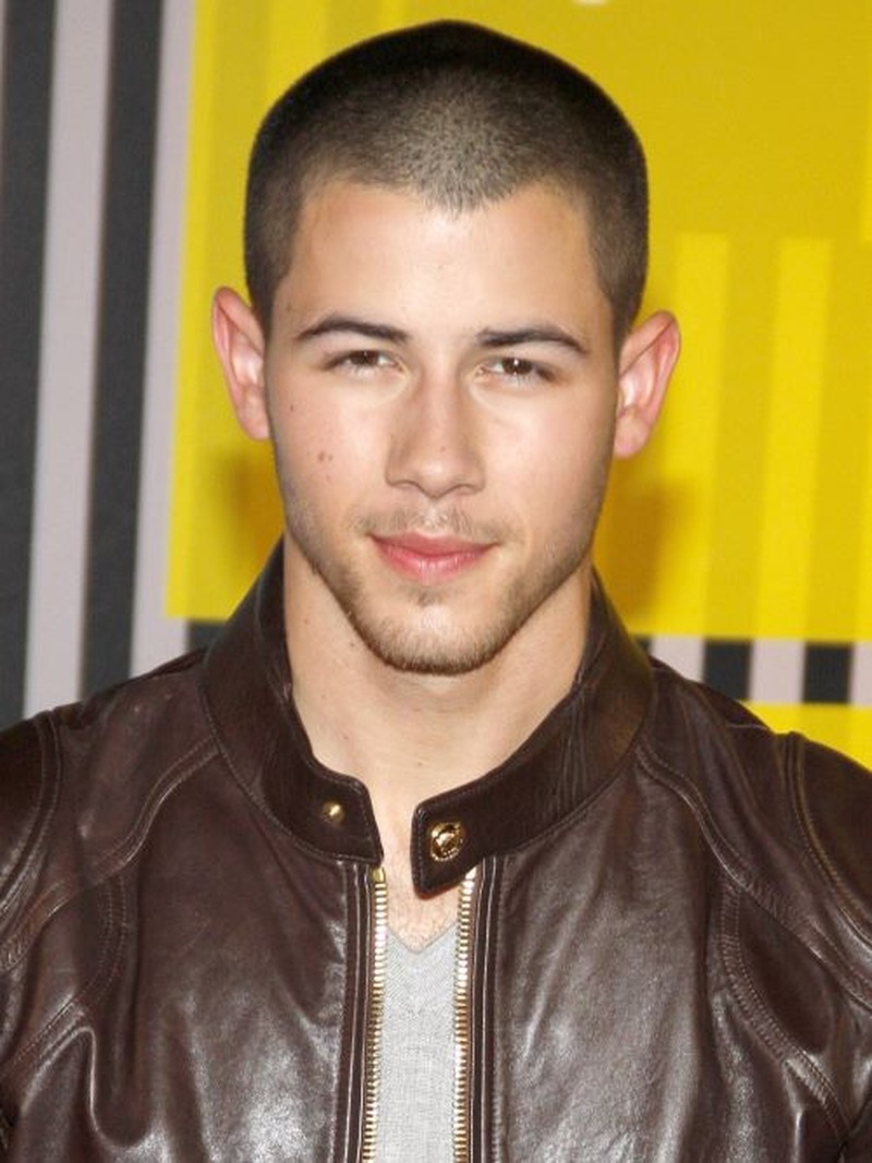 A man with a closely shaved induction buzz cut (very short hair, uniform length) and a clean, sharp appearance, wearing a brown leather jacket
