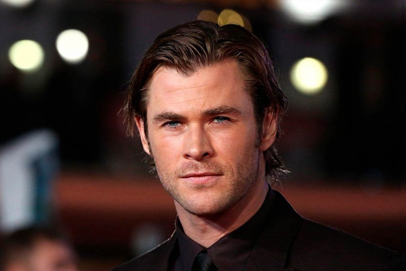 Chris Hemsworth sporting a medium-length wavy side-swept cut with textured layers and a polished finish, resembling his Thor Ragnarok hairstyle