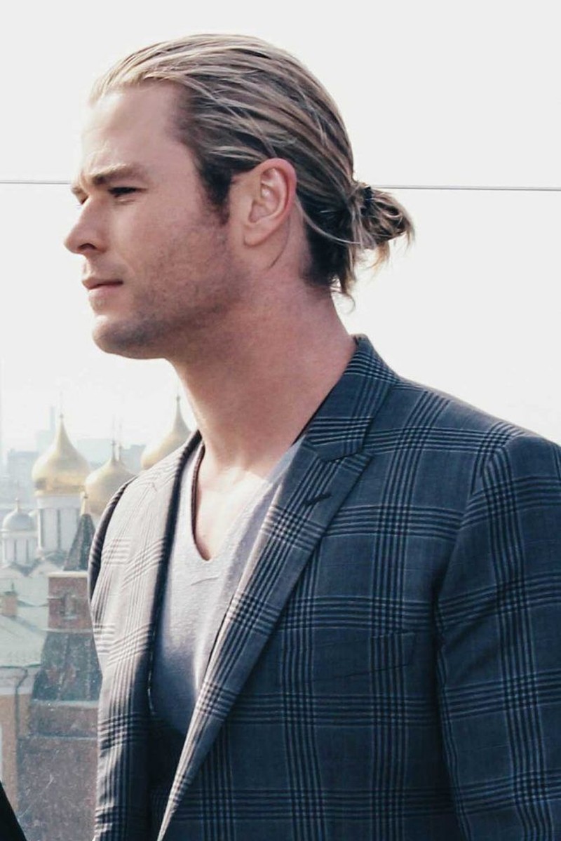 Chris Hemsworth with long wavy blonde hair styled in a low bun, featuring a textured slightly tousled cut with loose strands framing his face
