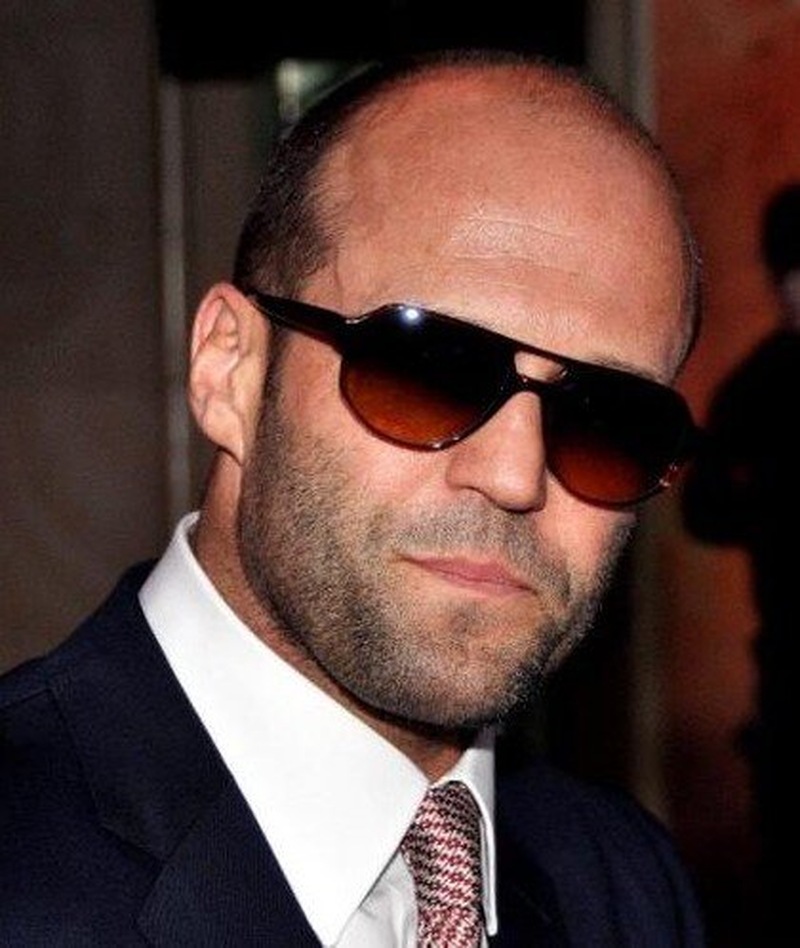 Jason Statham in a dark suit, white shirt, and patterned tie, showcasing his signature buzz cut and rugged beard style with sunglasses