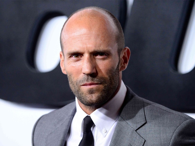Jason Statham in a sharp gray suit, showcasing a clean buzz cut that embraces his receding hairline, ideal for balding men seeking a stylish, confident look