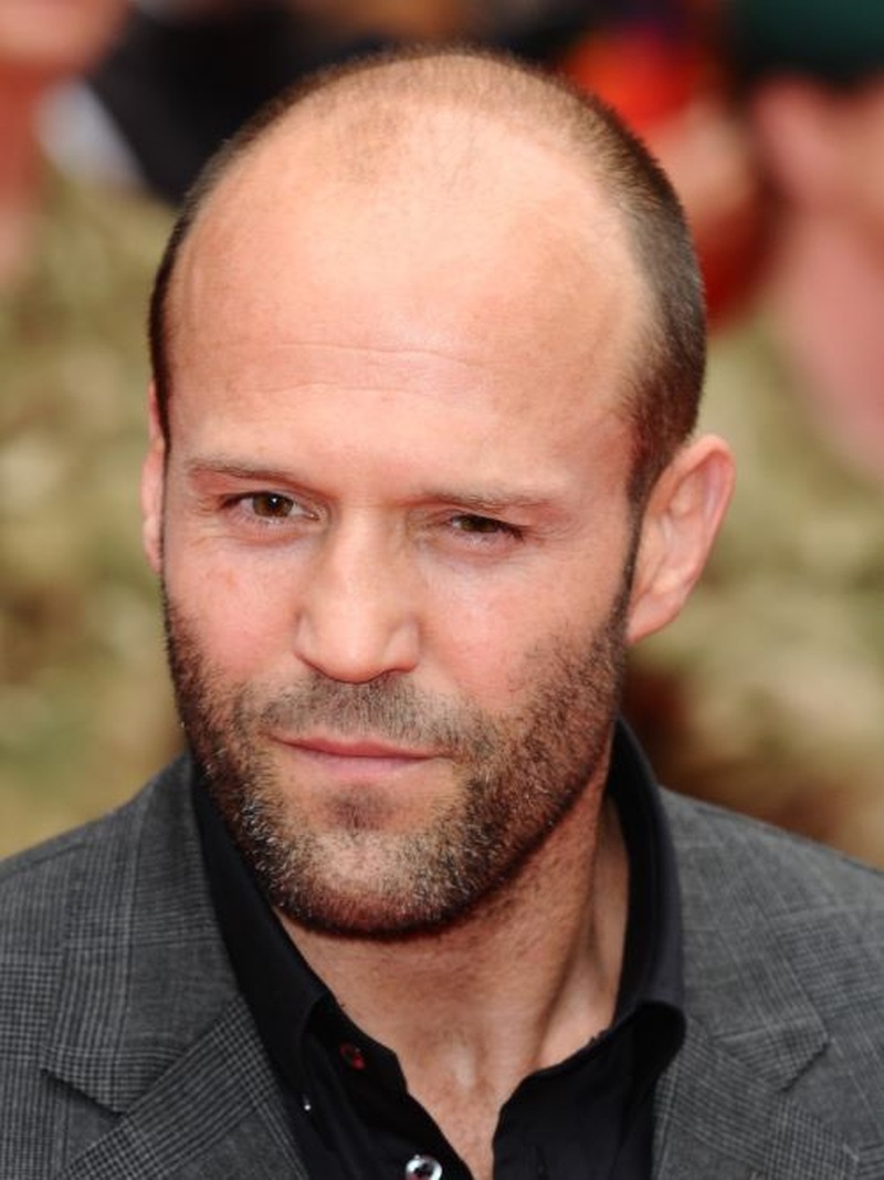 Jason Statham with a buzz cut, stubble beard, and sharp, rugged look in a gray blazer and black shirt