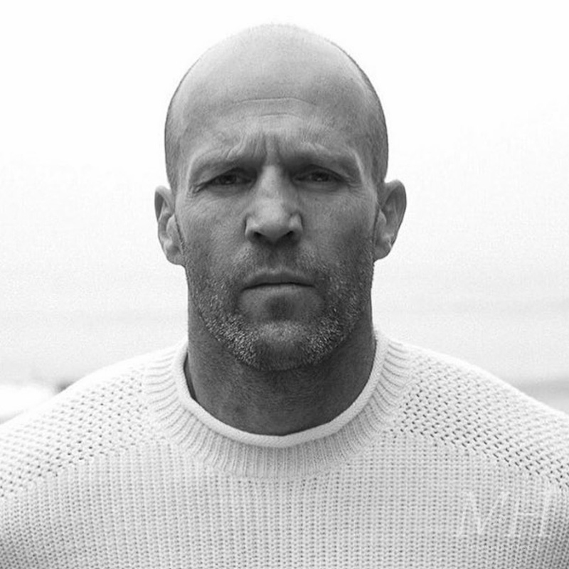 Jason Statham with a shaved buzz cut and short beard, wearing a knit sweater